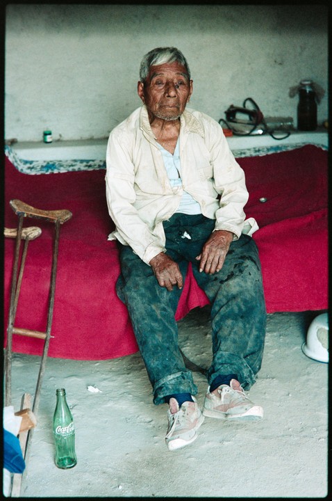 An elderly man with a weathered face sits on a red blanket, with a crutch and a glass Coca-Cola bottle nearby.
