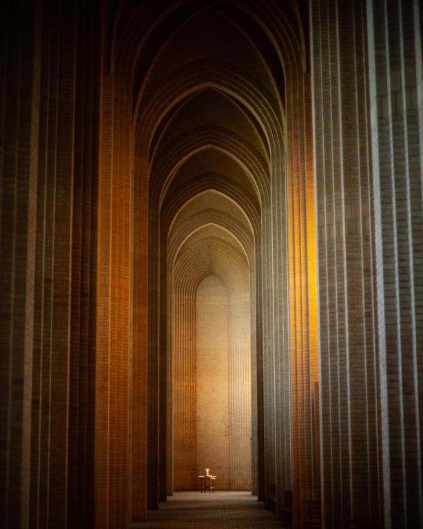 A long hallway with arched ceilings and columns leads to a small table with a lit candle.