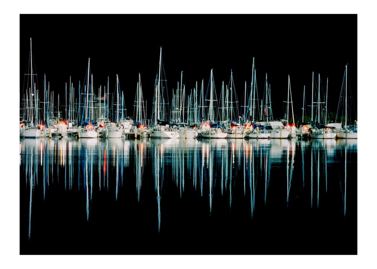 Many sailboats are docked at night, their masts and lights reflecting in the water.