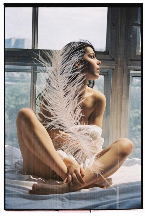 A woman sits on a bed by a window, holding a large feather.