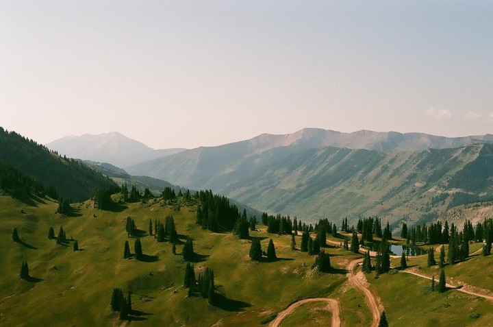 A winding dirt road leads through a green, tree-covered mountain landscape with a small pond and hazy mountains in the distance.