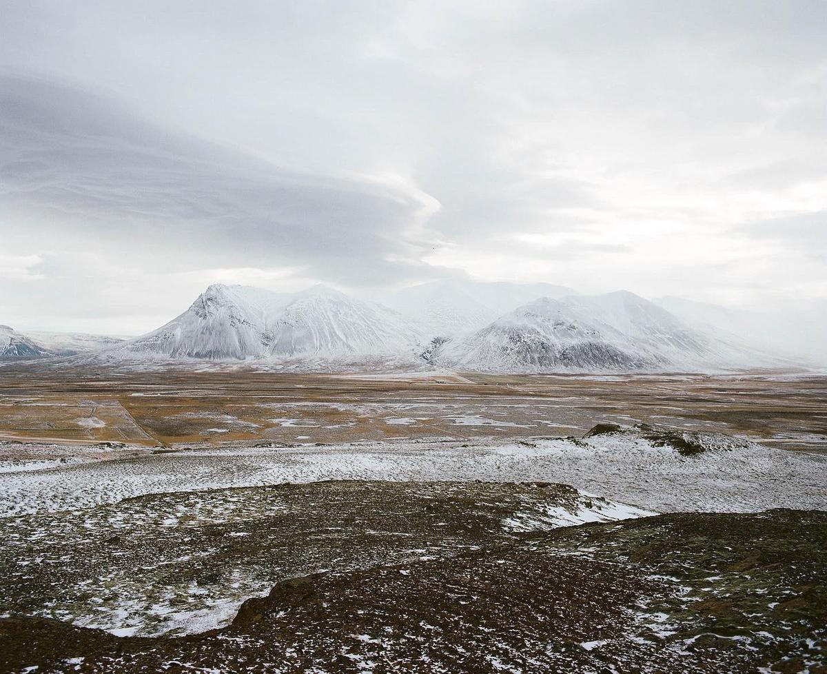 A vast, snow-dusted landscape stretches towards snow-capped mountains under a cloudy sky.