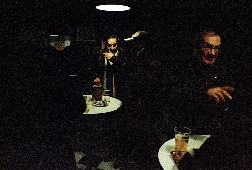 A man in sunglasses stands near a table with drinks in a dimly lit room while another man sits at a separate table with a drink.
