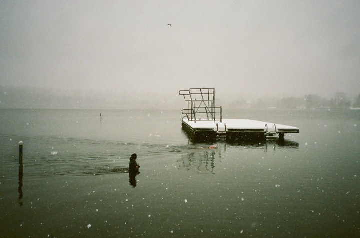 A person stands in a lake next to a snow-covered dock during a snowstorm.