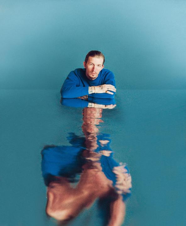 A man in a blue sweater is reflected in blue water.