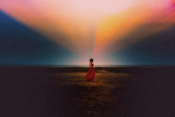 A woman in a red dress stands on a beach with a colorful sky above her.