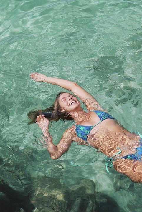 A woman in a blue bikini floats on her back in clear green water, laughing with her arms outstretched.