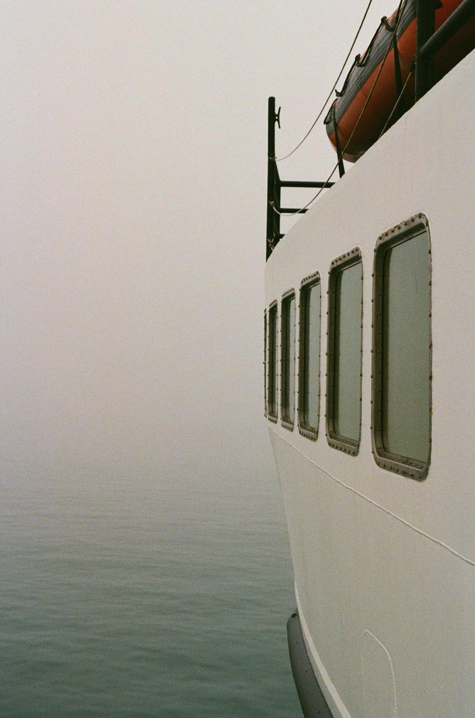 A white ferry boat with porthole windows cuts through foggy water.