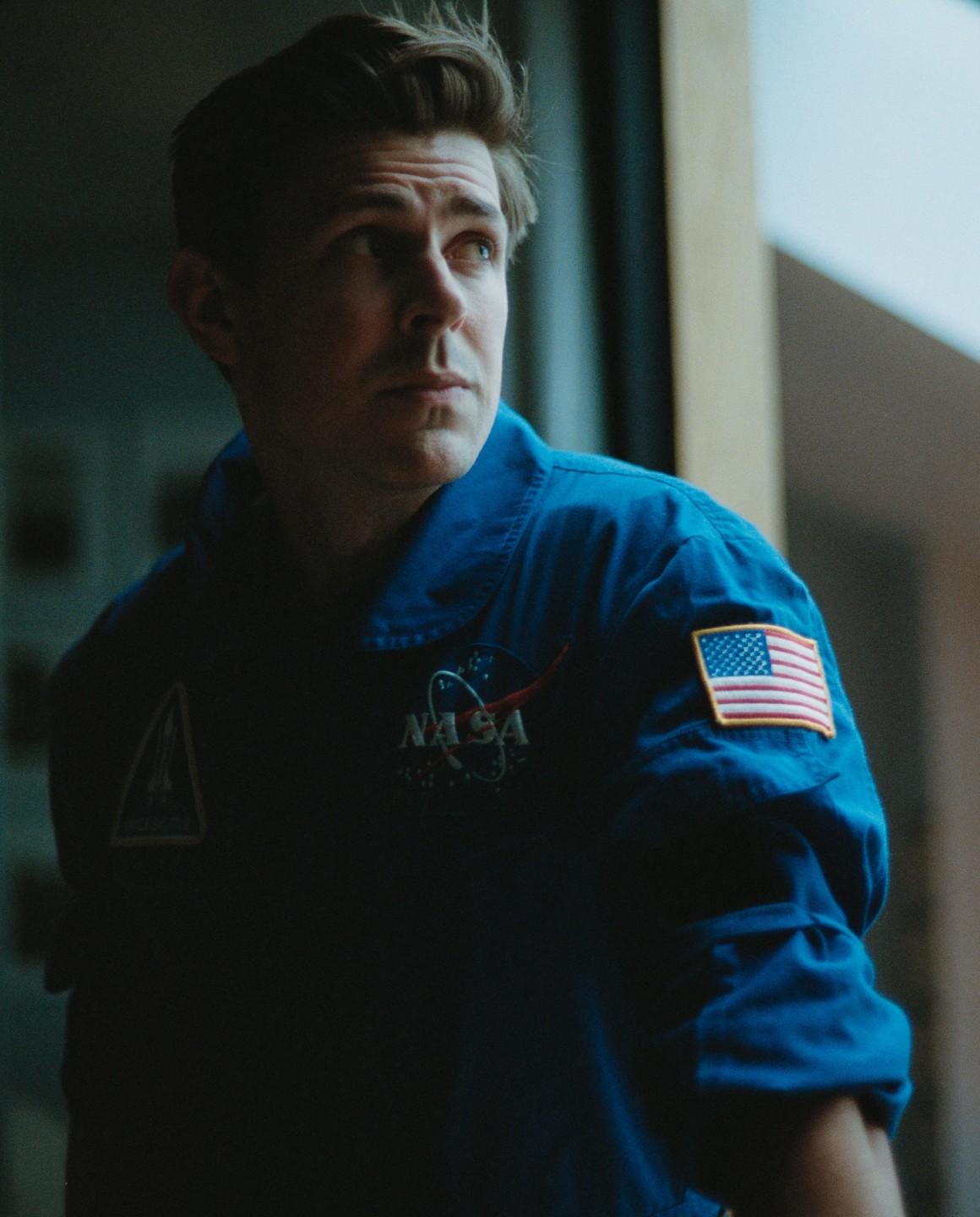 A man in a blue NASA flight suit looks up and to the right.