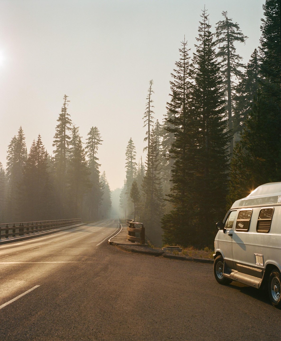 A white van is parked on the side of a road that leads into a hazy forest with tall pine trees.