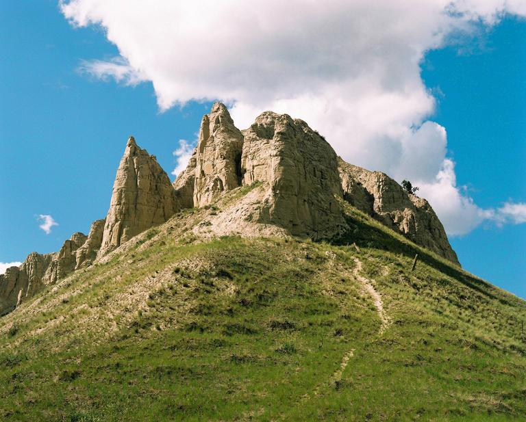 A grassy hill leads up to a rocky mountain peak under a blue sky with white clouds.