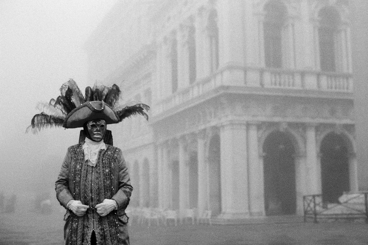 A person in a Venetian carnival costume with a feathered hat and mask stands in front of a building in the fog.