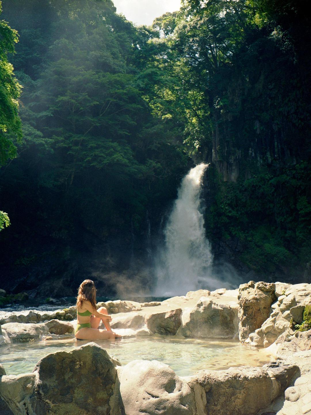 A woman sits in a hot spring with a waterfall and lush forest in the background.