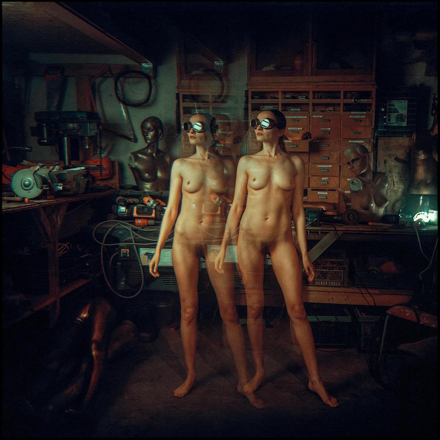 A nude woman with goggles stands in a workshop, with a faint double exposure of herself behind her.