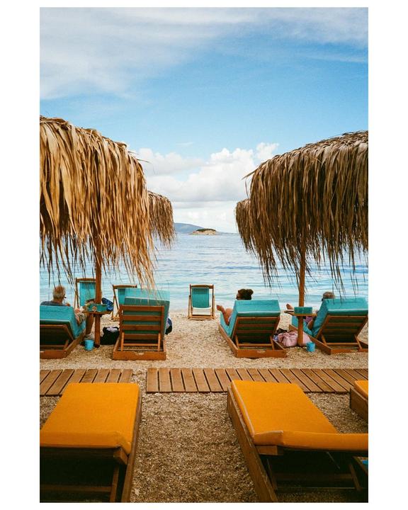 People relax on lounge chairs under straw umbrellas on a beach in Greece.