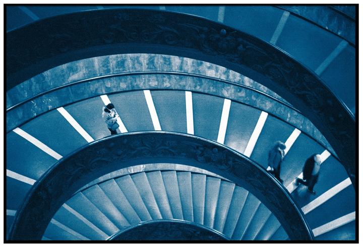 Three people walk down a spiral staircase with white stripes on the steps.