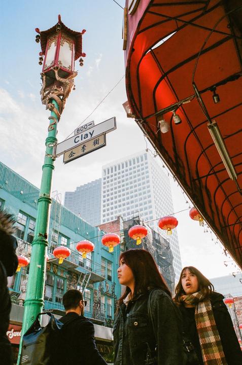 Two women and a man walk along a street in Chinatown, San Francisco, adorned with red lanterns and a decorative street lamp.
