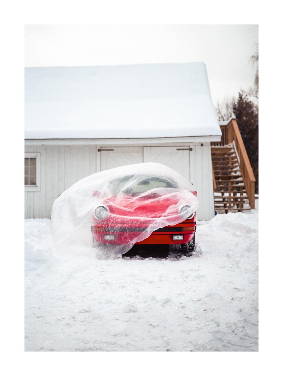 A red car covered with a plastic sheet is parked in the snow in front of a white building.