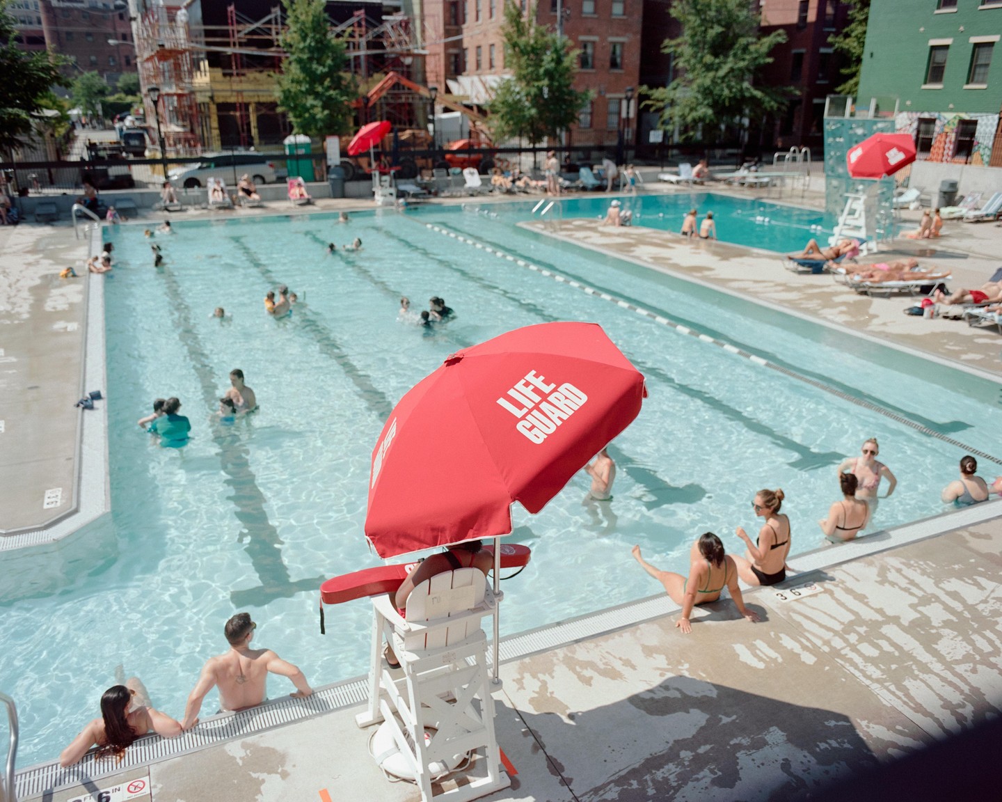 A lifeguard sits under a red umbrella next to a swimming pool filled with people.