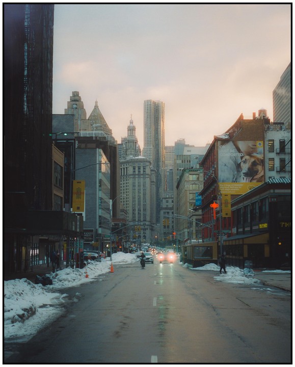 A street in a city is covered in snow and slush with cars driving down it and tall buildings on either side.