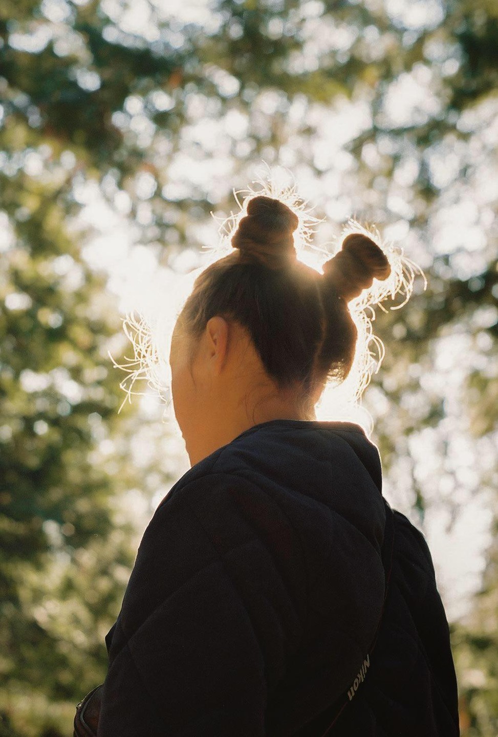 A woman with two buns in her hair is seen from behind, with sunlight backlighting her hair.