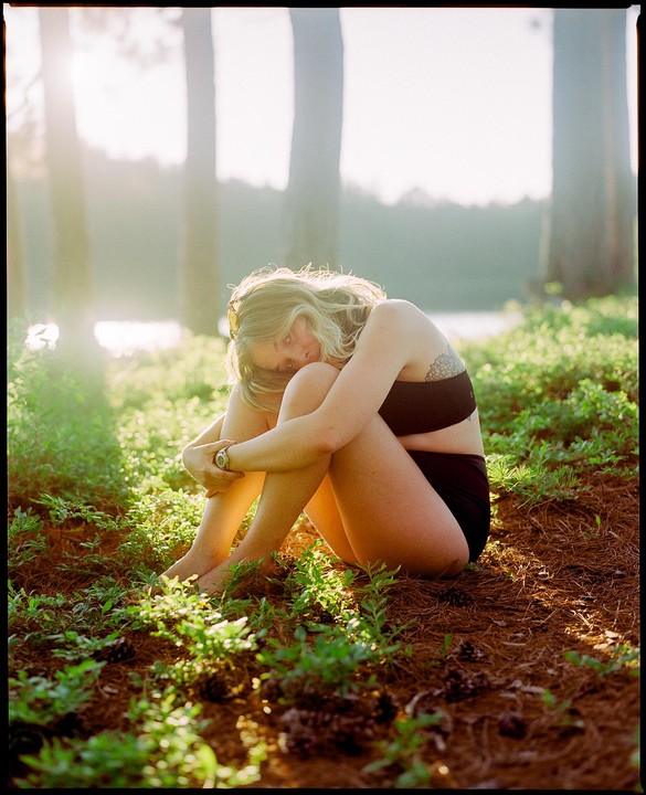 A woman sits on the ground in a forest with her knees pulled to her chest, looking towards the viewer.