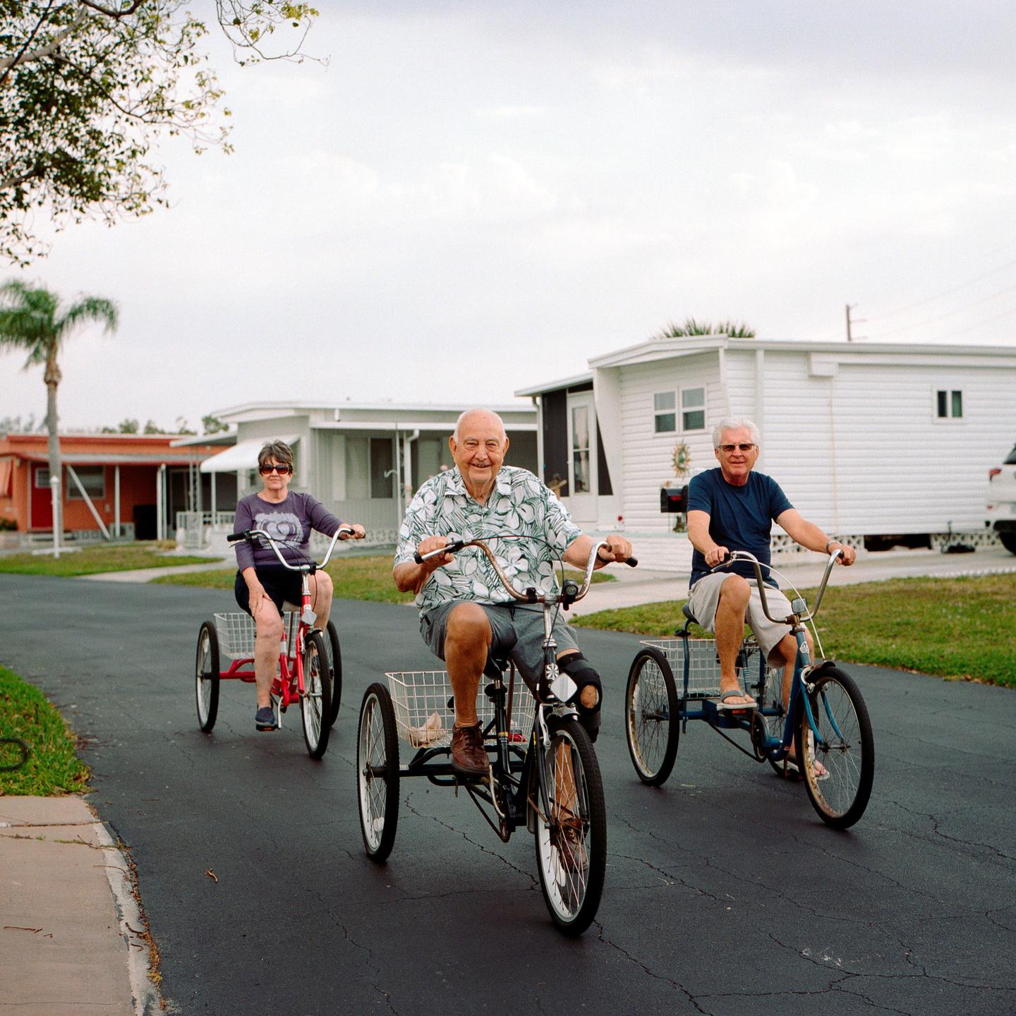 Three people ride tricycles down a paved road past mobile homes.