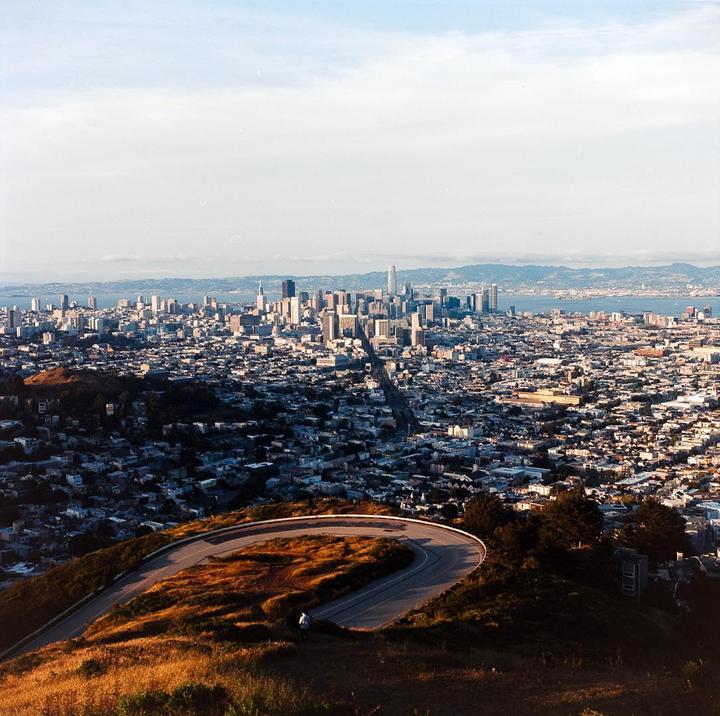 A winding road on a hill overlooks a city skyline and bay.