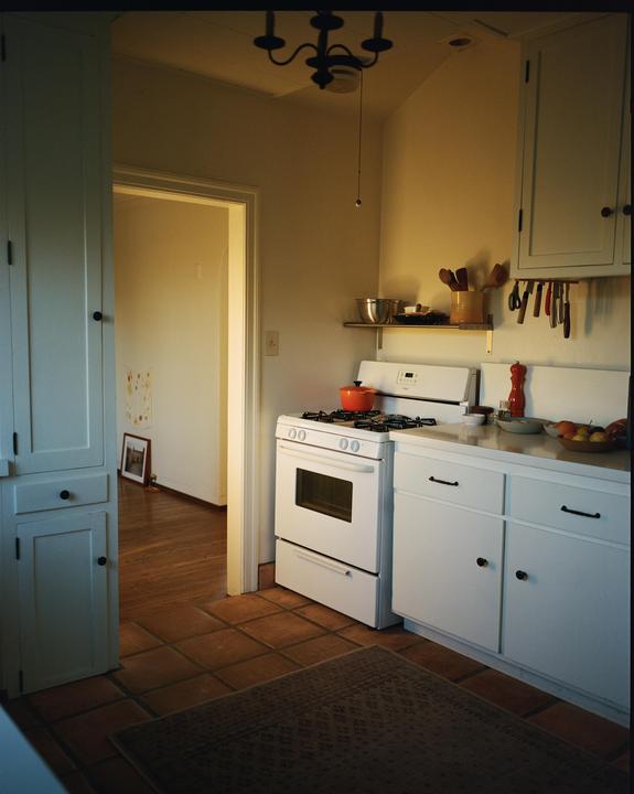 A kitchen with white cabinets and appliances is illuminated by warm light coming from a doorway.