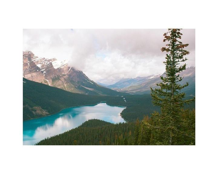 A turquoise lake is surrounded by a dense forest and snow-capped mountains under a cloudy sky.