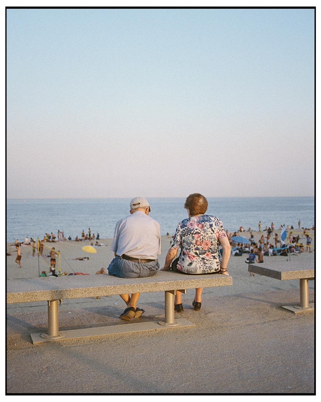 A couple sits on a bench overlooking a crowded beach with the ocean in the background.