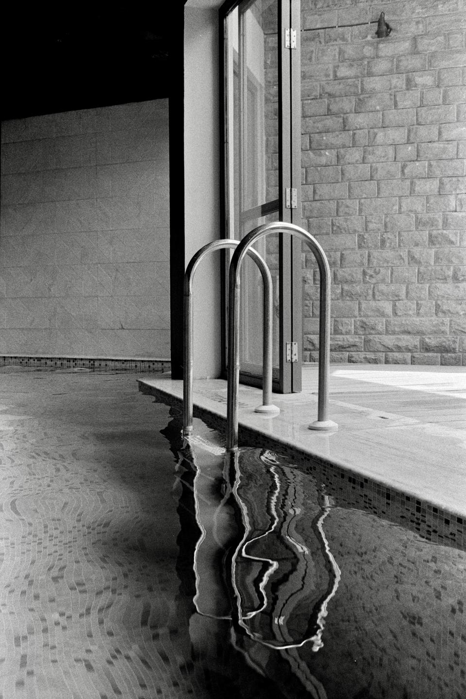 Two metal handrails extend into a swimming pool with a tiled edge, reflecting in the rippling water.