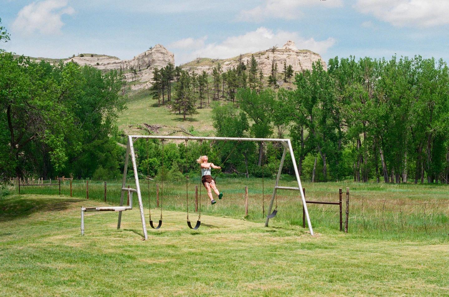 A woman swings on a swing set in a grassy field with trees and mountains in the background.