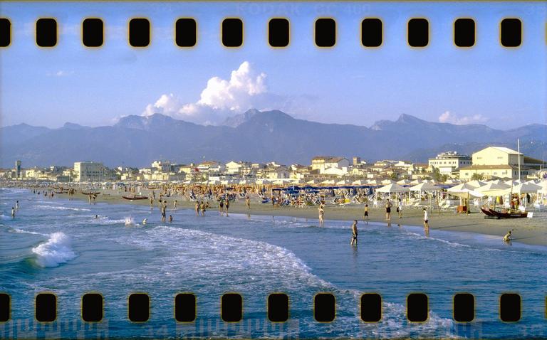 A wide shot shows a crowded beach with people in the water and on the sand, with a town and mountains in the background.
