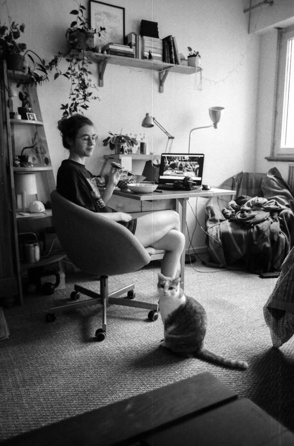 A woman sits at a desk with a laptop and a bowl of food, while a cat sits on the floor next to her.