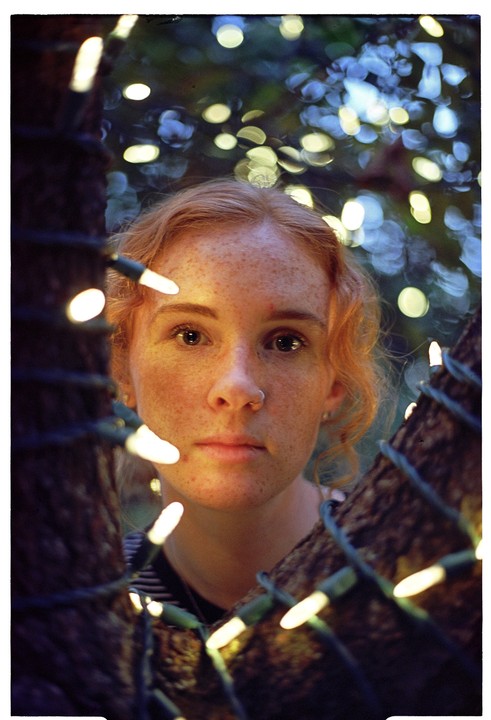 A woman with freckles and red hair looks directly at the viewer, framed by two tree trunks wrapped in string lights.