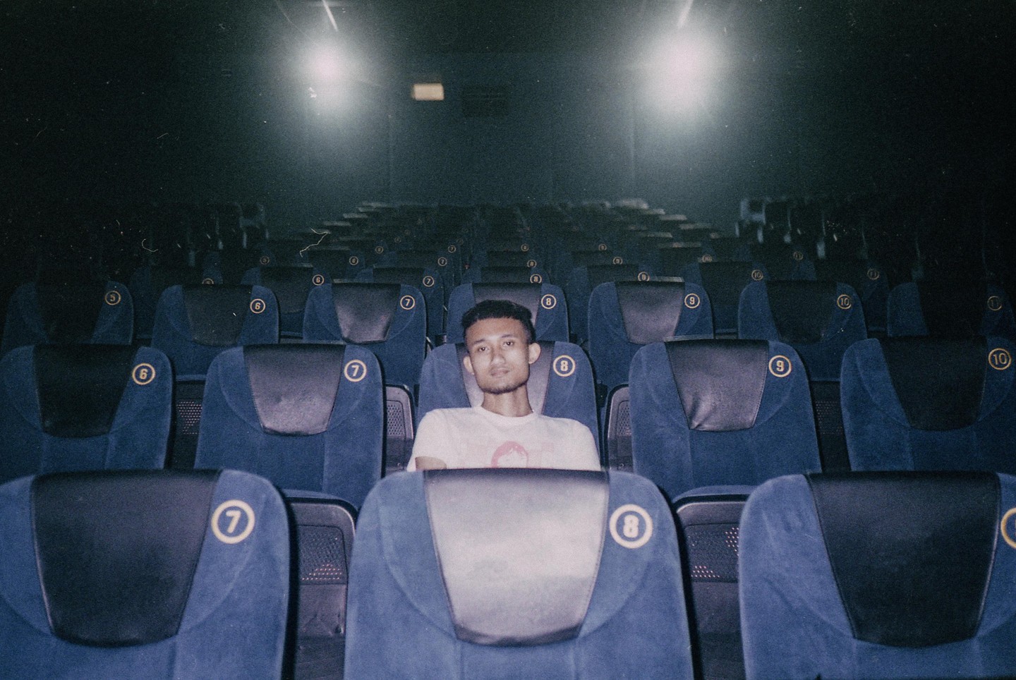 A man sits alone in a dark movie theater with rows of empty blue seats surrounding him.