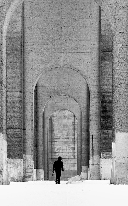 A man and a dog walk through a snowy arched passageway under the Hellgate Bridge.