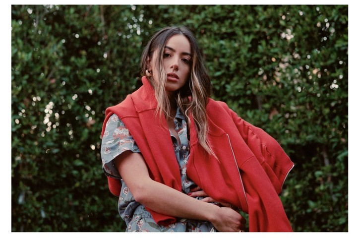 A woman with long hair and a red jacket over a patterned shirt poses in front of green foliage.