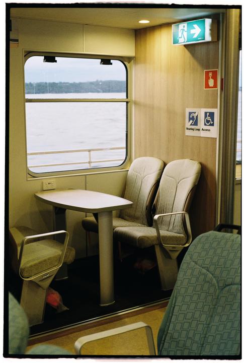 A ferry interior with a table and chairs is visible next to a window looking out onto water and a distant shoreline.