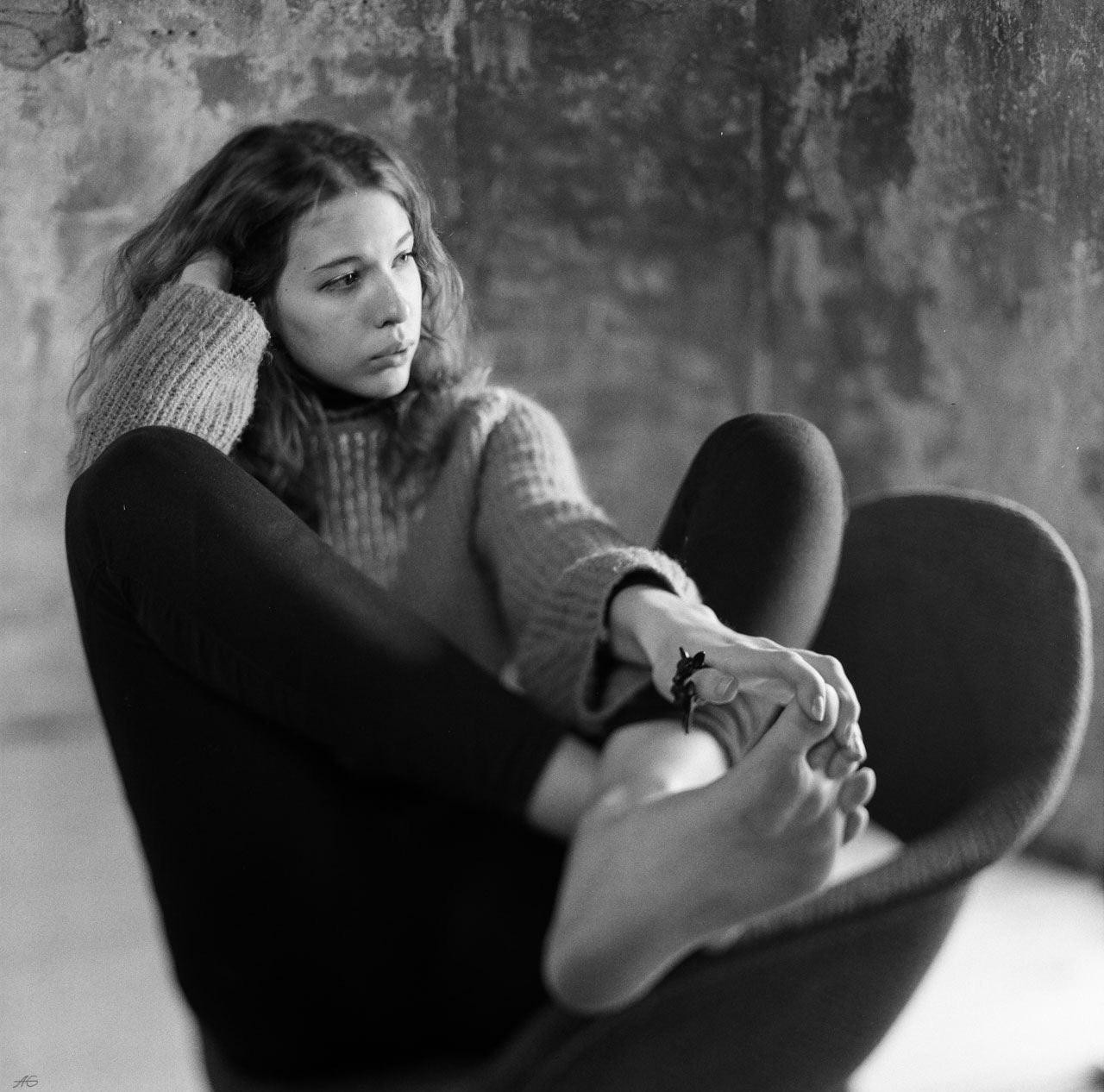 A young woman with curly hair sits in a chair, looking away with her hand on her head and her bare feet crossed.
