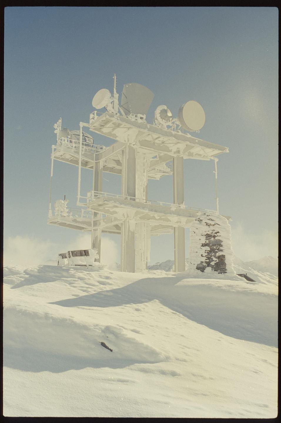 A snow-covered communication tower with multiple satellite dishes stands on a snowy mountain.