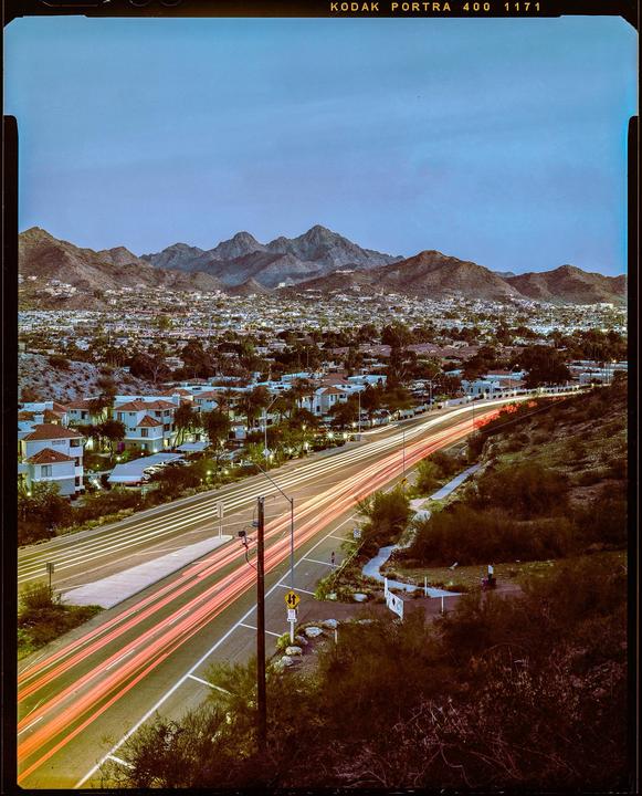 A long exposure captures light trails from cars on a road winding through a town nestled among mountains at dusk.