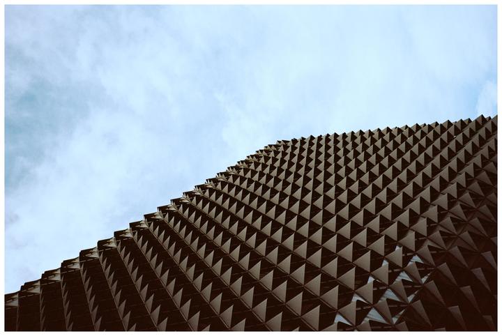 A building with a textured facade made of many small triangular shapes is shown against a cloudy sky.