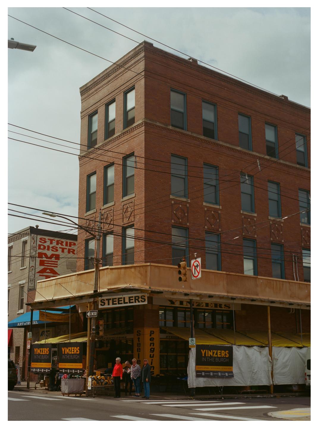 A group of people stand outside a brick building with a yellow awning and signs that say "Steelers" and "Yinzers in the Burgh" on a cloudy day.