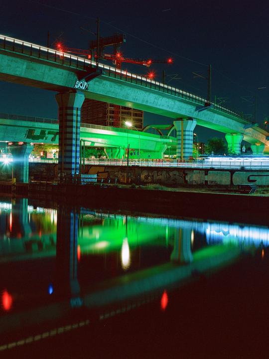 Light trails from vehicles on a bridge are reflected in the water below at night.