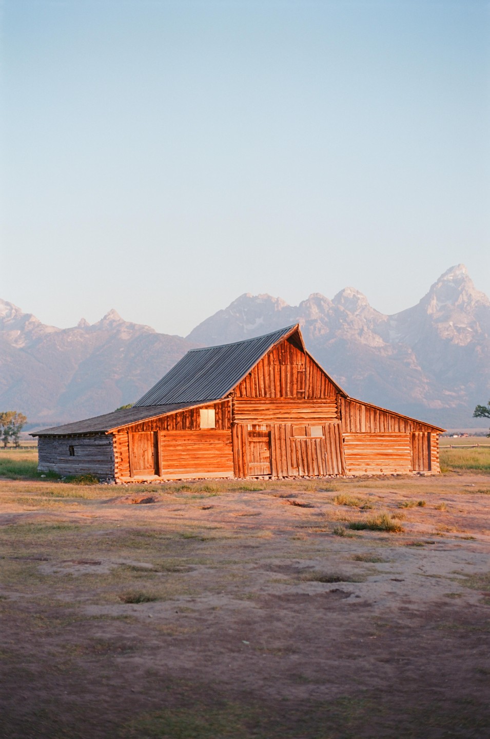 A wooden barn stands in a field with mountains in the background under a clear sky.