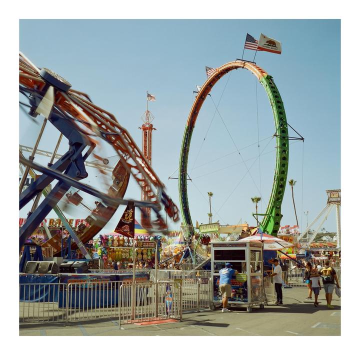 A fairground scene shows a large looping ride with an American flag and California flag at its peak, and another ride in motion in the foreground.