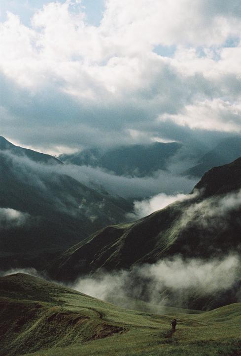A person walks on a grassy trail through a valley with mountains shrouded in mist and clouds.