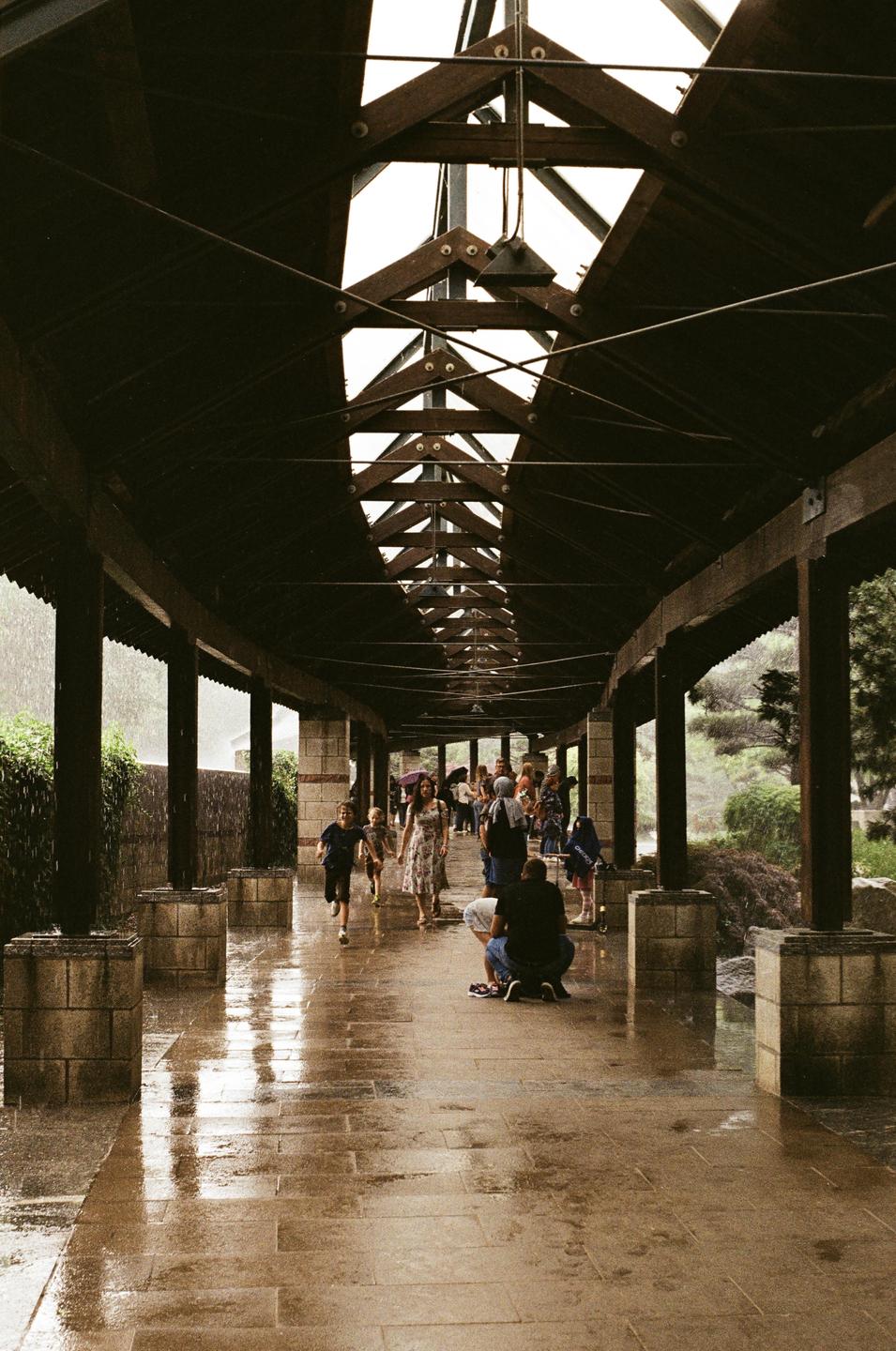 People walk and run on a wet stone path under a long wooden structure during a rain shower.